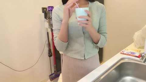 Media: Video of an Asian woman with straight black hair, wearing a light green cardigan and beige skirt, holding a cup, in a kitchen with a sink, vacuum cleaner, and cleaning supplies.