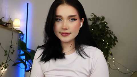 Media: Video of a young woman with fair skin, straight black hair, and light makeup, wearing white clothing, standing indoors with a green plant and fairy lights in the background.