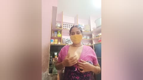 Media: Video of a woman with light brown skin and dark hair, wearing a yellow mask, a pink floral top, and a necklace, in a cluttered kitchen with pink walls and wooden shelves.