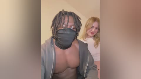 Media: Video of a shirtless Black man with dreadlocks and a black face mask, wearing a gray jacket, standing indoors with a blonde woman in a white top behind him.