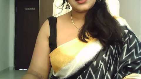 Media: Video of a woman with medium brown skin, wearing a black and white patterned sari with a mustard yellow border. She has long black hair and is indoors, possibly in a medical setting, as a \"Vaccination\" sign is visible in the background.