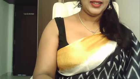 Media: Video of a South Asian woman with medium-brown skin, wearing a black sleeveless top with a white geometric pattern, and a large white and golden-brown bag resembling a bread loaf slung over her shoulder. She has long, dark hair and is smiling.