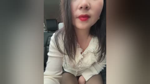 Media: A video of an Asian woman with long black hair, fair skin, and red lipstick, wearing a white lace blouse, seated in a car with black leather seats.