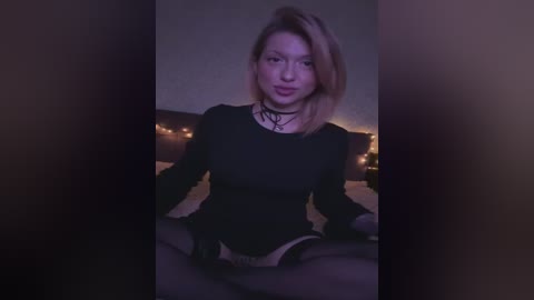 Media: Video of a light-skinned, blonde woman with a slender build, wearing a black choker and long-sleeve top, seated cross-legged on a bed with dimly lit string lights in the background.