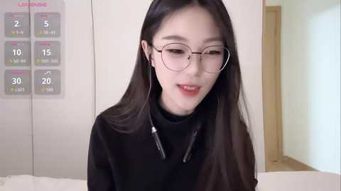 Media: Video of an East Asian woman with long black hair, wearing glasses, a black turtleneck, and headphones, smiling softly. Background features a calendar and light-colored walls.