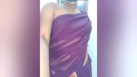 Media: Video of a woman wearing a purple saree with gold embroidery, partially covering her right breast, in a dimly lit room.