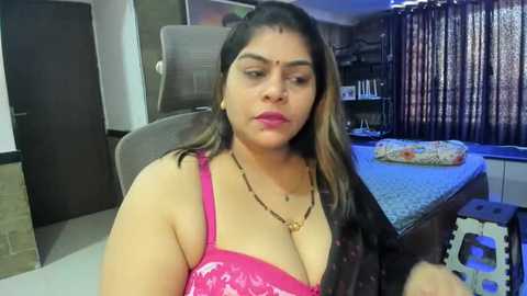 Media: Video of a plus-sized South Asian woman with long dark hair, wearing a bright pink sequin top, seated on a gray chair in a dimly lit room with a bed, curtains, and a door visible.