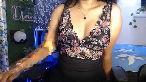 Media: Video of a woman with medium skin tone and long dark hair, wearing a black and pink floral lace top and high-waisted black skirt, standing in a room with blue floral wallpaper, green wall decor, and a chair.