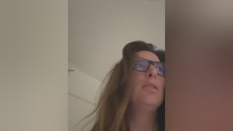 Media: A video of a young woman with long brown hair, wearing dark glasses and a casual top, standing in a dimly lit, plain room with white walls and a door.