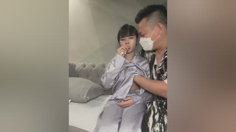 Media: Video: A young Asian boy in pajamas, coughing, is comforted by an adult male in a Hawaiian shirt and mask in a dimly lit room with a bed and pillows.