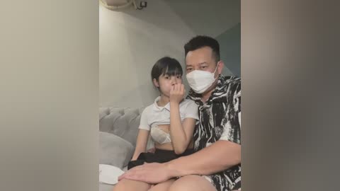 Media: A video of an Asian woman in a white top and bra, sitting on a couch, looking concerned, next to a man in a black shirt, wearing a mask, in a dimly-lit room.