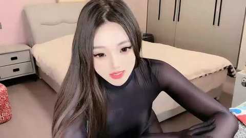 Media: Video of a young East Asian woman with long black hair, fair skin, and large eyes wearing a tight, black bodysuit. She sits on a bed in a modern bedroom with cream-colored bedding and light pink walls.