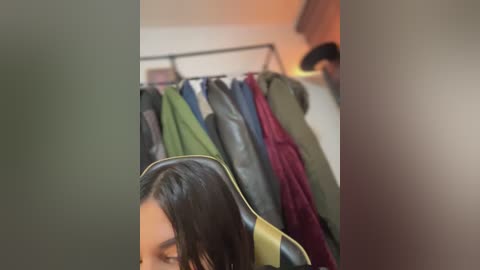 Media: Video of a woman with dark hair, wearing a black leather jacket, standing in a cluttered room with a coat rack displaying various colored coats.