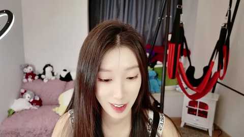 Media: A video of a smiling Asian woman with long, straight black hair, wearing a black and white patterned top, standing in a cluttered bedroom with a pink bed, stuffed toys, and workout equipment in the background.