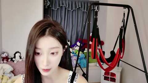 Media: Video of an East Asian woman with long brown hair, wearing a white bra, standing in a cluttered bedroom with a hanging swing, stuffed toys, and a window with blue curtains.