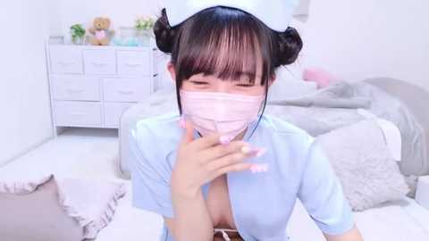 Media: Video of an Asian woman with black hair styled in buns, wearing a pink surgical mask, light blue nurse outfit, and hat, sitting on a white bed in a minimalist, clean bedroom.