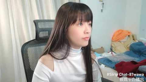 Media: Video of a young Asian woman with long, straight black hair and fair skin, wearing a white off-shoulder top, sitting on a black mesh chair in a messy room with clothes on a bed.