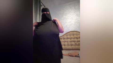 Media: A video of a person in a black mask, black shirt, standing in a dimly lit bedroom with a patterned wall, a headboard, and a bed with a quilt.