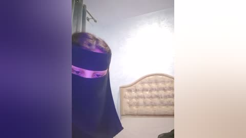 Media: Video of a woman in a black robe with a pink belt hanging from a hook in a dimly lit bedroom with a tufted headboard.