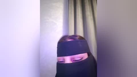 Media: Video of a person in a black burqa, face obscured, standing in front of a light gray curtain with a subtle purple hue. The image is slightly blurred, giving a sense of motion or low lighting.