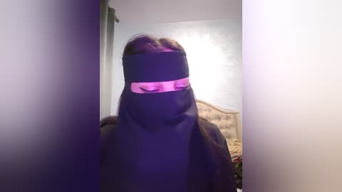 Media: A video of a person with long brown hair wearing a black blindfold, standing in a dimly lit room with a bed in the background.