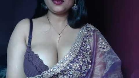 Media: A video of a South Asian woman with medium-dark skin, long black hair, and full lips, wearing a purple, lace-trimmed sari. She has large, dangling earrings and a delicate necklace. The background is a dark, muted purple.