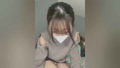 Media: Video of an East Asian woman with straight, dark hair and bangs, wearing a gray sweater with cut-out sleeves, a white mask, and seated on a bed with colorful bedding. The background is a plain, light-colored room.