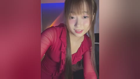 Media: A video of an East Asian woman with long, straight brown hair, wearing a red, ruffled blouse. She leans forward, smiling, with a soft, feminine glow. The background has a red and blue gradient wall.