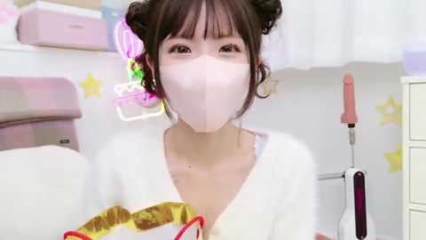 Media: Video of an East Asian woman with light skin, wearing a white mask, pink hair buns, and a white shirt with a yellow star pattern, in a pastel-colored room with a pink cabinet and a star-themed background.