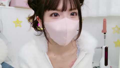 Media: A close-up video of a young woman with pale skin, wearing a pink face mask, white fur-trimmed jacket, and star-patterned earrings, in a clean, white room with a pink-handled hairdryer.