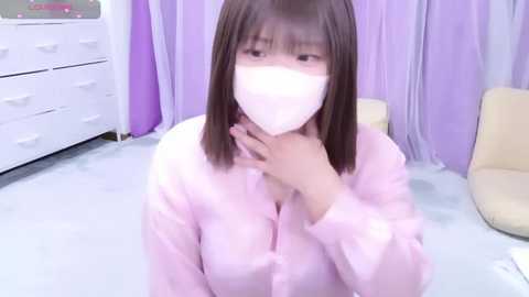 Media: Video of an Asian woman with auburn hair, wearing a pink button-up shirt and a white face mask, kneeling on a light blue floor in a room with pastel-colored walls and furniture.