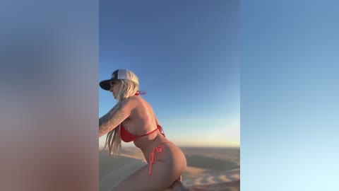 Media: Video of a blonde woman with tattoos in a red bikini, standing on sand dunes under a clear blue sky. She wears a white cap.