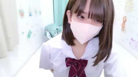 Media: Video of a young East Asian woman with straight, shoulder-length brown hair, wearing a white shirt, red plaid tie, and a white face mask in a brightly lit bathroom with pastel-colored wall art.