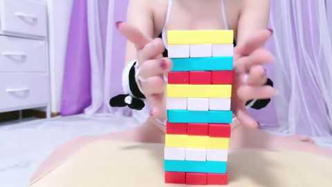 Media: Video of a person with fair skin, seated, wearing a white spaghetti-strap top, holding a brightly colored, stacked Jenga-like structure with fingers painted in red, yellow, and blue nail polish. Background features white furniture, light purple curtains, and a beige floor.