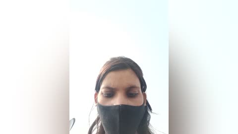 Media: A close-up video of a woman with long brown hair, wearing a black face mask, set against a stark white background. Her eyes are partially visible through the mask, creating a sense of mystery.