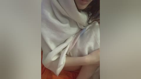 Media: A video shows a woman partially wrapped in a white towel, with her right arm and a glimpse of her dark hair visible. She wears orange shorts, and the background is blurred.