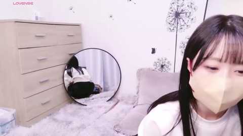 Media: Video of a young Asian woman with long black hair and pale skin, wearing a white mask, sitting on a fluffy rug in a minimalist bedroom with a beige dresser, a round mirror, and dandelion wall art.