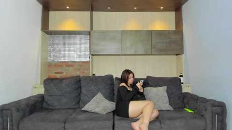 Media: Video of an Asian woman with straight dark hair, wearing a black off-shoulder dress, seated on a dark grey sectional sofa in a modern living room with wood paneling and recessed lighting.