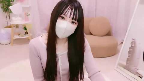 Media: Video of an Asian woman with long, straight black hair, wearing a light lavender cardigan and a white face mask, sitting in a pastel pink room with a beige armchair and a potted plant.