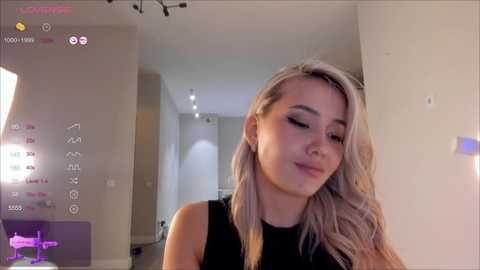 Media: A video of a young, fair-skinned, blonde woman with wavy hair, wearing a black top, standing in a modern, minimalist room with white walls and recessed lighting.