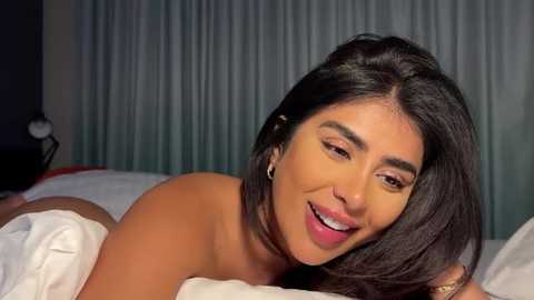 Media: Video of a topless woman with medium brown skin, long dark hair, and a cheerful expression lying on a white bed, against a muted blue curtain backdrop.