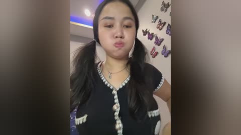 Media: Video of a young Asian woman with long black hair in pigtails, wearing a black dress with white trim, blowing a kiss in a room decorated with butterflies on the wall.