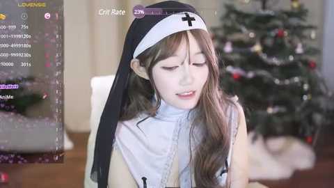 Media: Video of a young Asian woman dressed as a nun, sitting in a dimly lit room with a Christmas tree in the background. She has long, wavy brown hair and wears a white habit with a black cross.