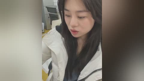 Media: A video of a young Asian woman with long black hair, wearing a white jacket, sitting indoors near a desk and chair. She has a neutral expression.