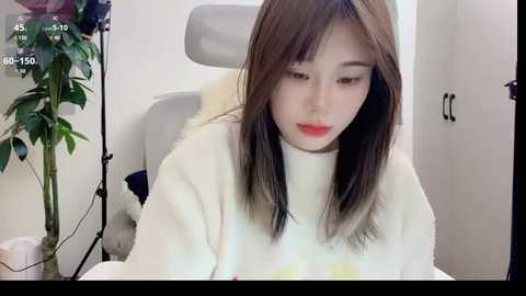 Media: Video of a young Asian woman with straight, shoulder-length brown hair, wearing a white turtleneck sweater, sitting at a desk, looking down. Background includes a gray office chair, a green plant, and a white wall.