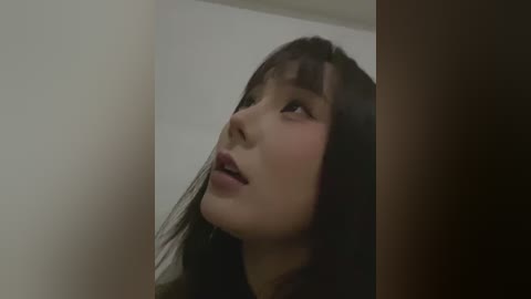 Media: A close-up video of an Asian woman with long black hair and fair skin, gazing upwards, likely indoors. She wears a dark top, and the background is minimalistic, featuring a beige wall and a wooden door frame.