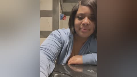 Media: Video of a woman with medium skin tone and shoulder-length black hair, wearing a light blue cardigan, leaning on a black marble counter in a modern bathroom.