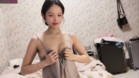 Media: A video of an Asian woman with dark hair, fair skin, and small breasts, wearing a grey camisole, standing in a cluttered bedroom with floral wallpaper.