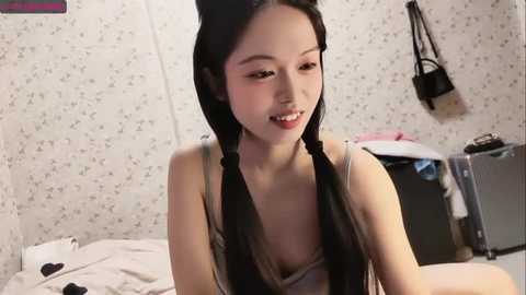 Media: A video of an Asian woman with long black hair in twin braids, smiling, wearing a tank top, in a cluttered bedroom with floral wallpaper and a black handbag hanging on the wall.