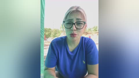 Media: Video of a woman with light skin, light blonde hair, and wearing black-rimmed glasses, a blue T-shirt, and a frown, standing outdoors in a blurry, green and blue-tinted background.